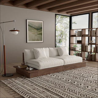 A modern, low-profile modular sofa with a dark wood frame is positioned in a cozy living room. The thick cushions and scatter pillows are upholstered in a light beige fabric. An attached side table on the left holds books and headphones, and a patterned area rug sits on the wooden floor underneath. Large windows, a framed botanical print, and a bookshelf complete the background.