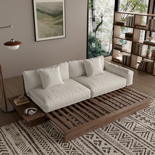 A modern, low-profile modular sofa bed with a dark wood frame is shown partially extended in a cozy living room. The thick cushions and scatter pillows are upholstered in a light beige fabric, resting on the visible slatted wood base. An attached side table on the left holds books and headphones.