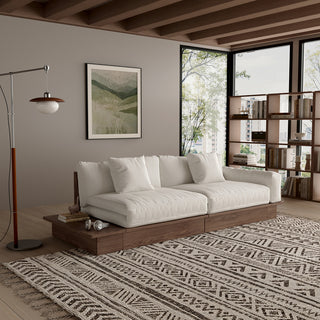A modern, low-profile modular sofa with a dark wood frame is positioned in a cozy living room. The thick cushions and scatter pillows are upholstered in a light beige fabric. An attached side table holds books and headphones, and a patterned area rug sits on the wooden floor underneath. Large windows, a framed botanical print, and a bookshelf complete the background.