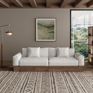 A modern, low-profile modular sofa with a dark wood base is positioned centrally against a taupe wall. The thick seat cushions, backrest pillows, and throw pillows are upholstered in a light cream fabric. A patterned area rug covers the floor in front, while a framed botanical art print, a floor lamp, and a window complete the living room setting.