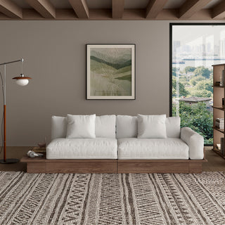 A modern, low-profile modular sofa with a dark wood frame is positioned centrally against a taupe wall. The thick seat cushions, backrest pillows, and throw pillows are upholstered in a light cream fabric. An extended wooden platform juts out from the left side of the sofa base. A patterned area rug covers the floor in front, while a framed botanical art print, a floor lamp, and a window complete the living room setting.