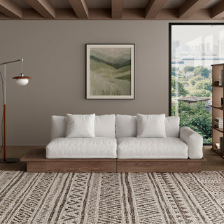A modern, low-profile modular sofa with a dark wood frame is positioned centrally against a taupe wall. The thick seat cushions, backrest pillows, and throw pillows are upholstered in a light cream fabric. An extended wooden platform juts out from the left side of the sofa base. A patterned area rug covers the floor in front, while a framed botanical art print, a floor lamp, and a bookshelf complete the living room setting.