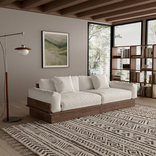 A modern, low-profile modular sofa with a dark wood frame is positioned in a cozy living room. The thick cushions and scatter pillows are upholstered in a light beige fabric. A patterned area rug sits on the wooden floor underneath, while a floor lamp, framed botanical print, and bookcase complete the background.