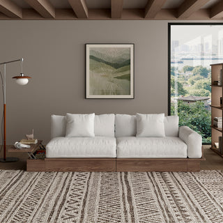 A modern, low-profile modular sofa with a dark wood frame is positioned against a taupe wall. The thick cushions and scatter pillows are upholstered in a light cream fabric. An attached side table on the left features a built-in compartment holding books and headphones. A patterned area rug sits on the floor in front, while a framed botanical art print, floor lamp, and window complete the living room.