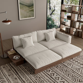 A modern, low-profile modular sofa bed with a dark wood frame is shown partially extended in a cozy living room. The thick cushions and scatter pillows are upholstered in a light beige fabric. An attached side table holds books and headphones, while a patterned area rug sits on the floor underneath. A framed botanical art print hangs on the neutral wall, and a tall wooden bookshelf filled with books stands in the background.