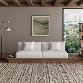 A modern low-profile sofa with white cushions sits on a wooden platform base in a bright, minimalist living room. The couch is adorned with small accent pillows and positioned in front of a neutral-toned wall decorated with a framed landscape artwork. A patterned area rug covers the floor in front of the sofa, while a large window to the right offers a view of a lush outdoor landscape.