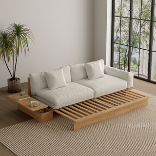 A wide, low-profile modular sofa bed with a natural wood frame is shown partially extended to reveal its wooden slat base. The thick cushions and scatter pillows are upholstered in a light beige fabric. An attached side table features a built-in drawer holding small items, with books and headphones resting on top. A potted palm tree stands beside the sofa in a room with a large window and a textured beige area rug.