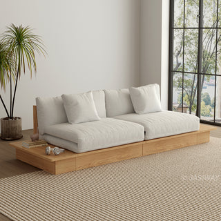 A wide, low-profile modular sofa with a natural wood frame sits in a bright room next to a large window. The thick cushions and scatter pillows are upholstered in a light beige fabric. An extended wooden platform functions as a side table, holding small books and headphones. A potted palm tree stands beside the sofa on a textured beige area rug.