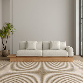 A modern, low-profile modular sofa with a natural wood base is positioned against a neutral grey wall. The thick cushions and scatter pillows are upholstered in a light cream fabric. An extended wooden platform juts out from the left side of the sofa base. A potted palm plant stands to the left, and a textured beige area rug covers the floor in front.