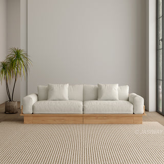 A modern, low-profile modular sofa with a natural wood base is positioned centrally against a neutral grey wall. The thick seat cushions, armrests, and back cushions are upholstered in a light cream fabric, complete with matching scatter pillows. A potted palm plant stands to the left, and a textured beige area rug covers the floor in front.