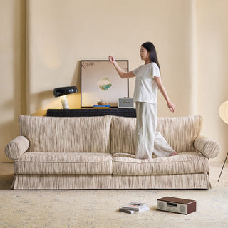 JASIWAY textured linen blend sofa, featuring a minimalist upholstery with a subtle linear weave, highlighting its plush, oversized bolsters and the architectural solid wood side panels, perfect for contemporary living rooms.-Front view with model stepping on the sofa