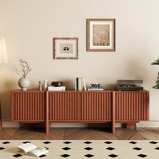 A modern wooden media console with vertical slat paneling is set against a warm beige wall. The top of the cabinet is decorated with books, vases, a small bowl, and a record player. Two framed abstract artworks hang on the wall above the unit. In the foreground, a patterned area rug rests on a herringbone wood floor, completing the cozy living space.