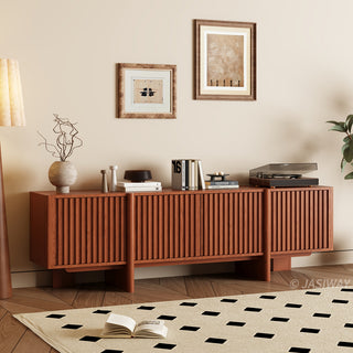 A modern reddish-brown wooden media console with vertical slat paneling is set against a warm beige wall. The top of the cabinet is decorated with books, vases, a small bowl, and a record player. Two framed abstract artworks hang on the wall above the unit. In the foreground, a patterned area rug rests on a herringbone wood floor, completing the cozy living space.