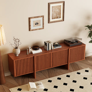 A modern reddish-brown wooden media console with vertical slat paneling is set against a warm beige wall. The top of the cabinet is decorated with books, vases, a small bowl, and a record player. Two framed abstract artworks hang on the wall above the unit. In the foreground, a patterned area rug rests on a herringbone wood floor, completing the cozy living space.