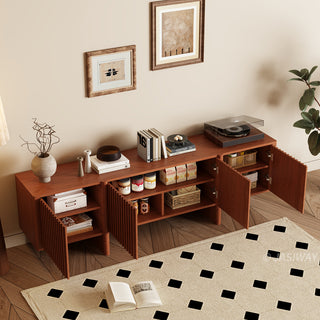 A modern reddish-brown wooden media console is set against a warm beige wall with two framed art prints hanging above. The cabinet doors are open, revealing organized storage compartments filled with jars, boxes, and baskets. The surface of the unit is decorated with books, vases, and a record player on the right. In the foreground, a patterned area rug with black diamond shapes lies on a herringbone wooden floor.