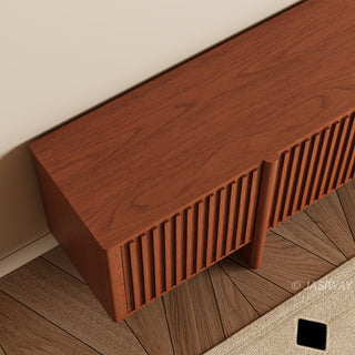 A close-up, high-angle shot captures a portion of a modern reddish-brown wooden media console. The cabinet features distinctive vertical slatted paneling and sits against a warm beige wall. Below the console, a patterned rug rests on a herringbone wooden floor.