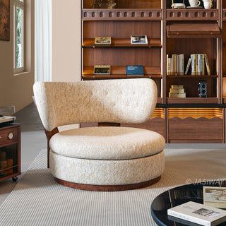 A modern, rounded armchair upholstered in a textured cream and gold fabric sits centered on a light-colored rug. The chair features a thick, circular seat cushion and a curved, button-tufted backrest supported by a dark wood frame. In the background, a large wooden bookshelf filled with books and decorative items adds warmth to the room.