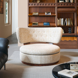 A modern, rounded armchair upholstered in a textured cream and gold fabric is set on a light-colored rug. The chair features a thick, circular seat cushion and a curved, button-tufted backrest with wooden supports. In the background, a large wooden bookshelf filled with books and decorative items is visible against the wall.