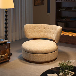 A modern, rounded armchair upholstered in a textured cream and gold fabric sits in a warm, dimly lit room. The chair features a thick, circular seat cushion and a curved, button-tufted backrest supported by a wooden base. A tall floor lamp stands to the left, while a dark wooden bookcase and a small cabinet with cans are visible in the background.