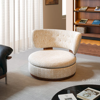 A modern, rounded armchair upholstered in a textured cream and gold fabric is set on a light-colored rug. The chair features a thick, circular seat cushion and a curved, button-tufted backrest with wooden support. In the background, white sheer curtains hang by a window alongside a wooden bookcase filled with books.