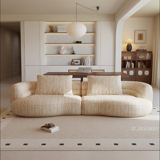 A beige textured modern curved sectional sofa featuring low-profile seating and matching back cushions, placed in a contemporary living room with a neutral area rug and built-in shelving.