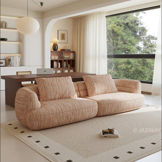 A beige textured modern curved sectional sofa featuring low-profile seating and matching back cushions, placed in a contemporary living room with a neutral area rug, a wooden dining table, and large windows.