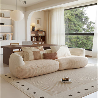 A beige textured modern curved sectional sofa featuring low-profile seating and matching back cushions, placed in a contemporary living room with a neutral area rug and built-in shelving.