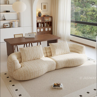 A beige textured modern curved sectional sofa featuring low-profile seating and matching back cushions, placed in a contemporary living room with a neutral area rug, a wooden dining table, and large windows.