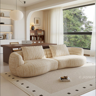 A beige textured modern curved sectional sofa featuring low-profile seating and matching back cushions, placed in a contemporary living room with a neutral area rug, a wooden dining table, and large windows.