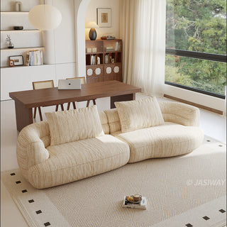 A beige textured modern curved sectional sofa featuring low-profile seating and matching back cushions, placed in a contemporary living room with a neutral area rug, a wooden dining table, and large windows.