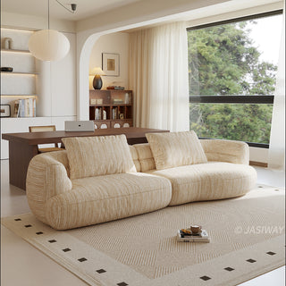A beige textured modern curved sectional sofa featuring low-profile seating and matching back cushions, placed in a contemporary living room with a neutral area rug, a wooden dining table, and large windows.