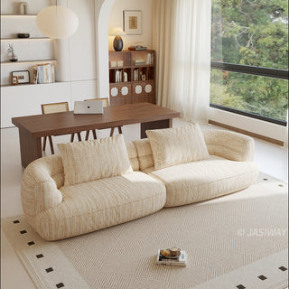 A beige textured modern curved sectional sofa featuring low-profile seating and matching back cushions, placed in a contemporary living room with a neutral area rug, a wooden dining table, and large windows.