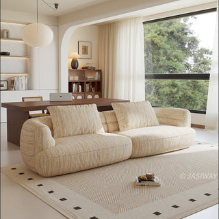 A beige textured modern curved sectional sofa featuring low-profile seating and matching back cushions, placed in a contemporary living room with a neutral area rug, a wooden dining table, and large windows.