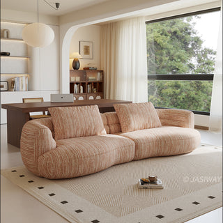 A beige textured modern curved sectional sofa featuring low-profile seating and matching back cushions, placed in a contemporary living room with a neutral area rug, a wooden dining table, and large windows.