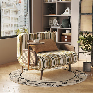 Modern oversized accent armchair with green and yellow striped fabric upholstery, solid wood legs, woven rattan armrests, and an integrated adjustable side table tray, placed on a patterned round rug in a sunlit reading nook.