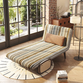 High-angle lifestyle shot of a convertible accent chair reclined into chaise lounge mode in olive green and cream striped bouclé fabric, featuring a brown lumbar pillow and woven rattan side panel, placed in a sunlit industrial loft near floor-to-ceiling windows.