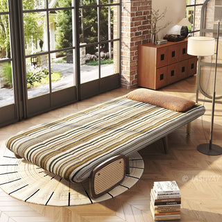 High-angle perspective view of a convertible accent chair fully opened into a single sleeper bed in olive green and brown striped bouclé fabric, featuring a brown pillow, woven rattan side panel, and supporting metal legs, styled in a sunlit industrial loft near French windows.