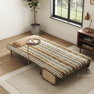 High-angle wide shot of a convertible sleeper accent chair fully flattened into a single sleeper bed in olive green and brown striped upholstery, featuring a built-in swiveling side tray table with a coffee cup, woven rattan armrest panel, and a pillow, styled in a modern bedroom setting near a window.
