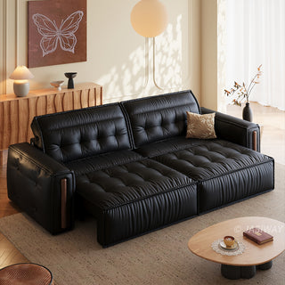 A modern black leather sofa is set in a bright, warmly lit living room. A small patterned pillow rests on the sofa, while a wooden coffee table sits on a rug in the foreground. In the background, a wooden console table is decorated with a butterfly art piece and a lamp.
