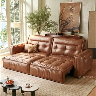 A modern, tufted brown leather sofa bed is positioned in a sunlit living room, partially extended into a chaise lounge configuration. It rests on a patterned rug alongside a unique, kidney-shaped two-tiered coffee table holding a bowl of fruit and a small dessert. The background features a large window, a potted tree, a wooden console table with decor, and a large framed floral artwork.