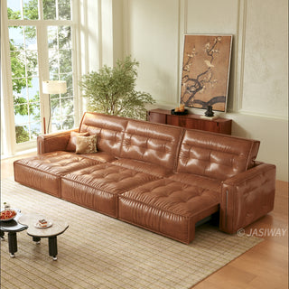 A modern, tufted brown leather sofa bed is positioned in a sunlit living room, partially extended into a chaise lounge configuration. It rests on a patterned rug alongside a unique, kidney-shaped two-tiered coffee table holding a bowl of fruit and a small dessert. The background features a large window, a potted tree, a wooden console table with decor, and a large framed floral artwork.