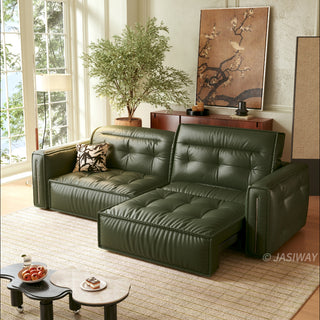 A modern, tufted dark green leather sofa bed is positioned in a sunlit living room, partially extended into a chaise lounge configuration. It rests on a patterned rug alongside a unique, kidney-shaped two-tiered coffee table holding a bowl of fruit and a small dessert. The background features a large window, a potted tree, a wooden console table with decor, and a large framed floral artwork.