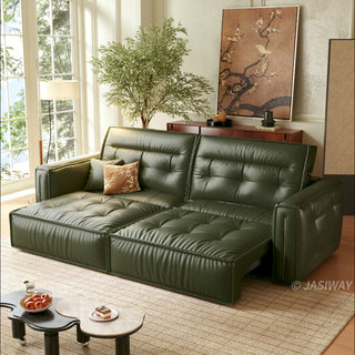 A modern, tufted dark green leather sofa bed is positioned in a sunlit living room, partially extended into a chaise lounge configuration. It sits on a patterned rug alongside a unique, kidney-shaped two-tiered coffee table holding a bowl of apples and a small dessert. The background features a large window, a potted tree, a wooden console table with decor, and a large piece of framed floral wall art. Natural light streams in from the left, illuminating the scene.
