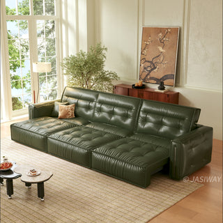 A modern, tufted dark green leather sofa bed is positioned in a sunlit living room, partially extended into a chaise lounge configuration. It rests on a patterned rug alongside a unique, kidney-shaped two-tiered coffee table holding a bowl of fruit and a small dessert. The background features a large window, a potted tree, a wooden console table with decor, and a large framed floral artwork.