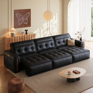 A spacious modern black leather sectional sofa is set in a warmly lit living room. The sofa features a tufted backrest and an extended seating area configured as a bed. An oval wooden coffee table sits on a large rug in the foreground, while a console table with a butterfly artwork decorates the background.