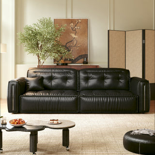 A modern, tufted black leather sofa is centered in a brightly lit, elegant living room. It rests on a textured light-colored rug alongside a uniquely shaped two-tiered coffee table holding a bowl of fruit and a small dessert. The background features a large potted tree, a framed floral wall art piece, and a folding screen.