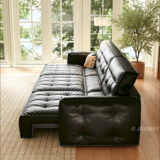 A black leather sofa bed is fully extended into a flat bed position in a bright, sunlit room. It is placed on a light-colored rug against a backdrop of large windows with a view of greenery. A potted plant and a floor lamp stand near the window, completing the cozy scene.