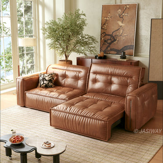 A modern, tufted brown leather sofa bed is positioned in a sunlit living room, partially extended into a chaise lounge configuration. It rests on a patterned rug alongside a unique, kidney-shaped two-tiered coffee table holding a bowl of fruit and a small dessert. The background features a large window, a potted tree, a wooden console table with decor, and a large framed floral artwork.