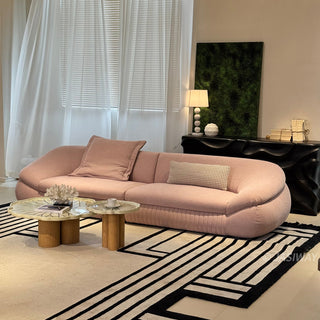 RINGCHEN modern curved cloud sofa featuring premium blush textured upholstery, highlighted by its signature sculptural low-profile silhouette, channel-tufted base detailing, and deep-seated organic cushioning for a high-end minimalist living room aesthetic.-Three quarter view with warm lighting