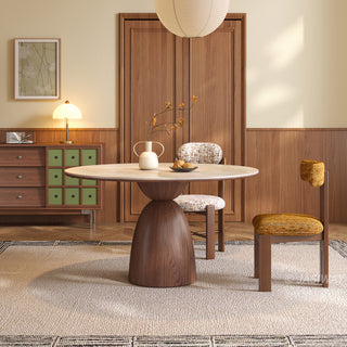 This elegant dining room features a round stone-top table supported by a distinct wooden pedestal base. A white vase holding delicate branches and a small dish rest on the table surface. Chairs with patterned upholstery are arranged around the table, set against a wood-paneled wall and a patterned rug on the floor.